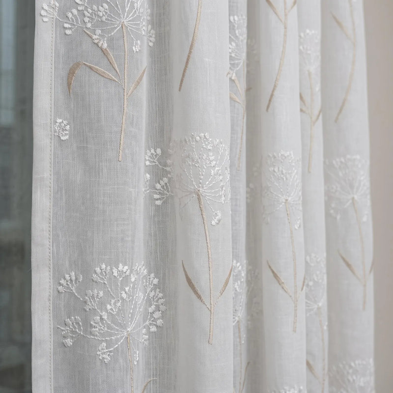 White sheer curtain fabric with embroidered dandelion and wildflower motifs in white and light tan, shown hanging in soft folds.